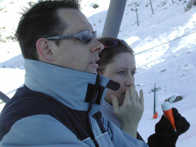 Two people in winter clothing on a ski lift; one applies lip balm while the other looks ahead.