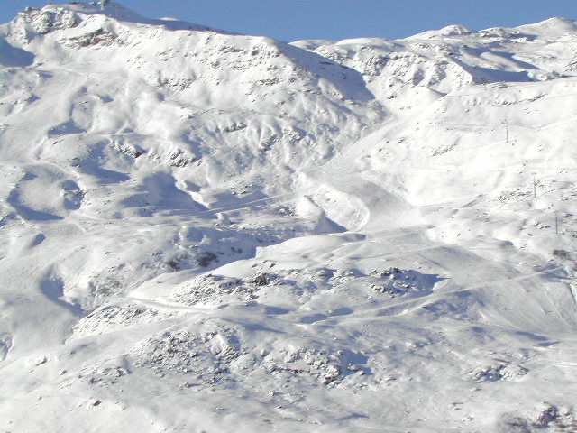 Snow-covered mountain slopes with ski trails winding down the terrain under a clear blue sky.