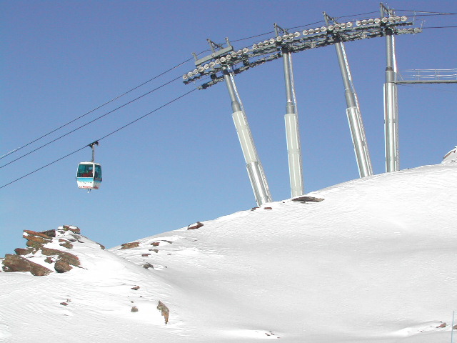 A cable car moves over a snowy mountain slope, supported by tall metal structures under a clear blue sky.