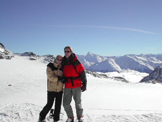 Two people in winter jackets stand on a snowy mountain, smiling and posing for a photo.