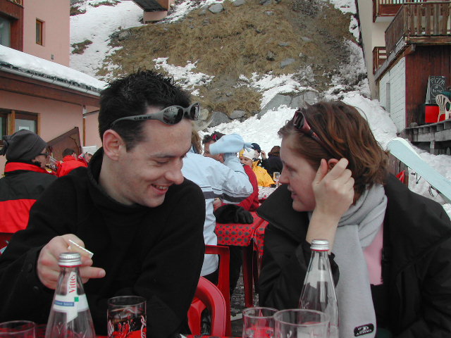 A man and woman sit at an outdoor table, talking and smiling, with drinks and bottles in front of them.