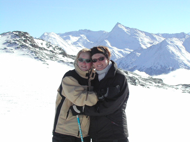 Two people in winter jackets and sunglasses hug and smile on a snowy mountain with peaks in the background.