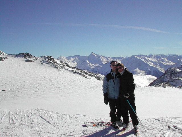 Two people in ski gear stand close together on a snowy mountain with skis, smiling at the camera.