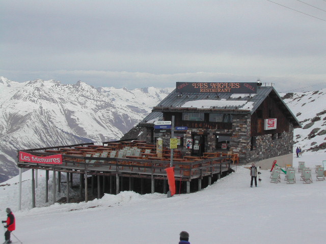 A stone restaurant with a wooden terrace in a snowy mountain setting, with skiers nearby at Les Menuires.