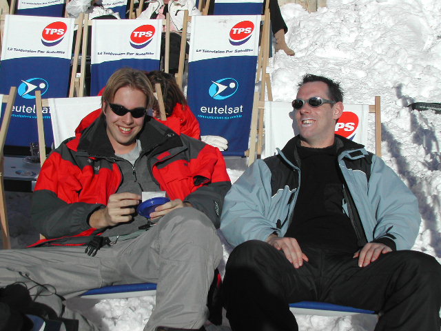 Two men in winter jackets and sunglasses relax on lounge chairs in the snow, smiling and enjoying the sun.