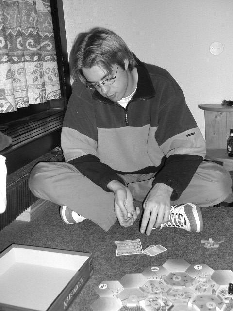 A person sits on the floor, rolling dice while playing a board game with hexagonal tiles and cards.