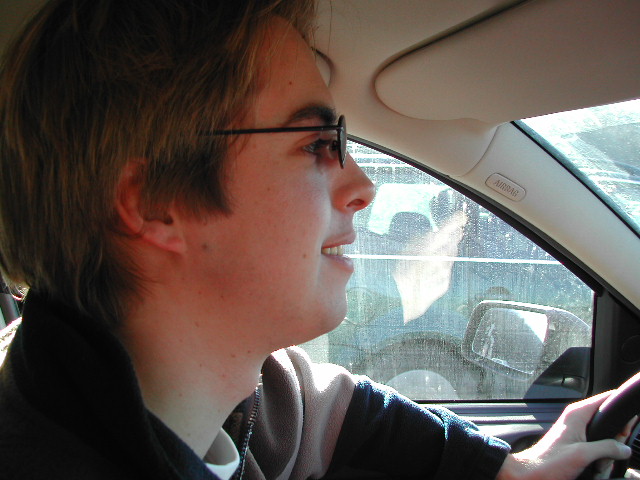 A person wearing glasses is driving a car, looking ahead with a slight smile.