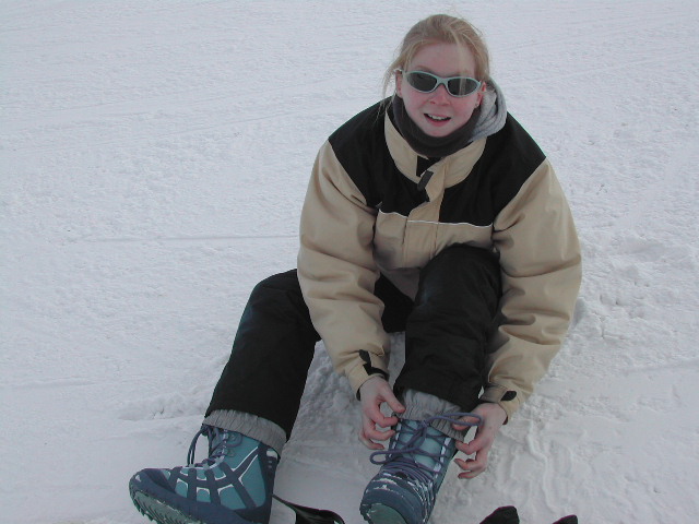 A person sitting on the snow, wearing winter gear, tying their boot laces while looking at the camera.