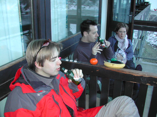 Three people sit on a balcony, drinking beer and eating snacks.