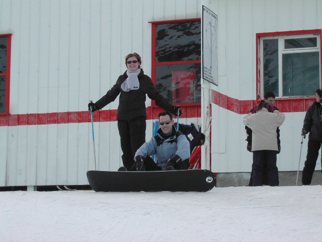 A person stands on skis behind another sitting on a snowboard, both smiling in front of a snowy building.