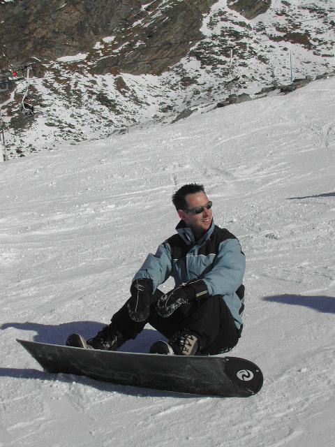 A person in a winter jacket and gloves sits on the snow with a snowboard, smiling and looking sideways.