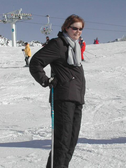 A person in a black ski outfit and sunglasses stands on a snowy slope, holding a ski pole.