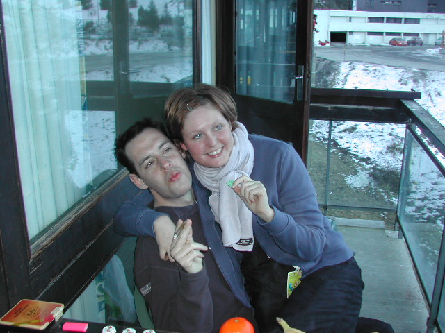 A man and woman sit closely on a balcony, smiling and holding cigarettes, with a snowy landscape behind them.