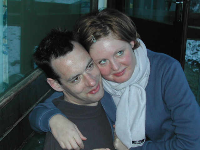 A woman in a blue sweater hugs a man, both smiling and looking off to the side.
