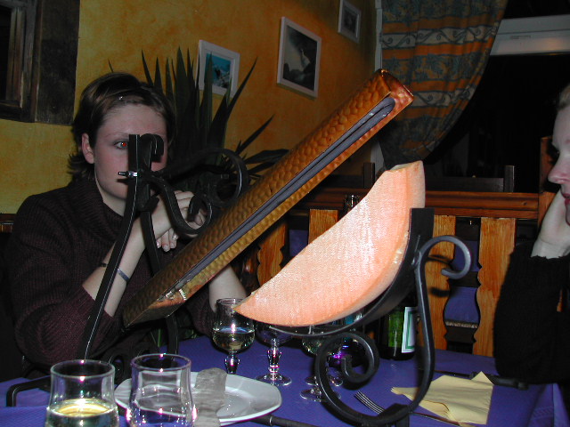 A person sits at a table with a large wedge of cheese melting under a raclette grill.