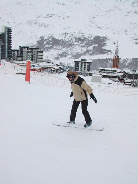 A person snowboarding down a snowy slope, wearing a beige and black jacket, goggles, and gloves.