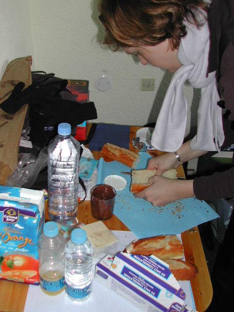 A person wearing a scarf prepares a sandwich on a cluttered table with bread, water bottles, and food items.