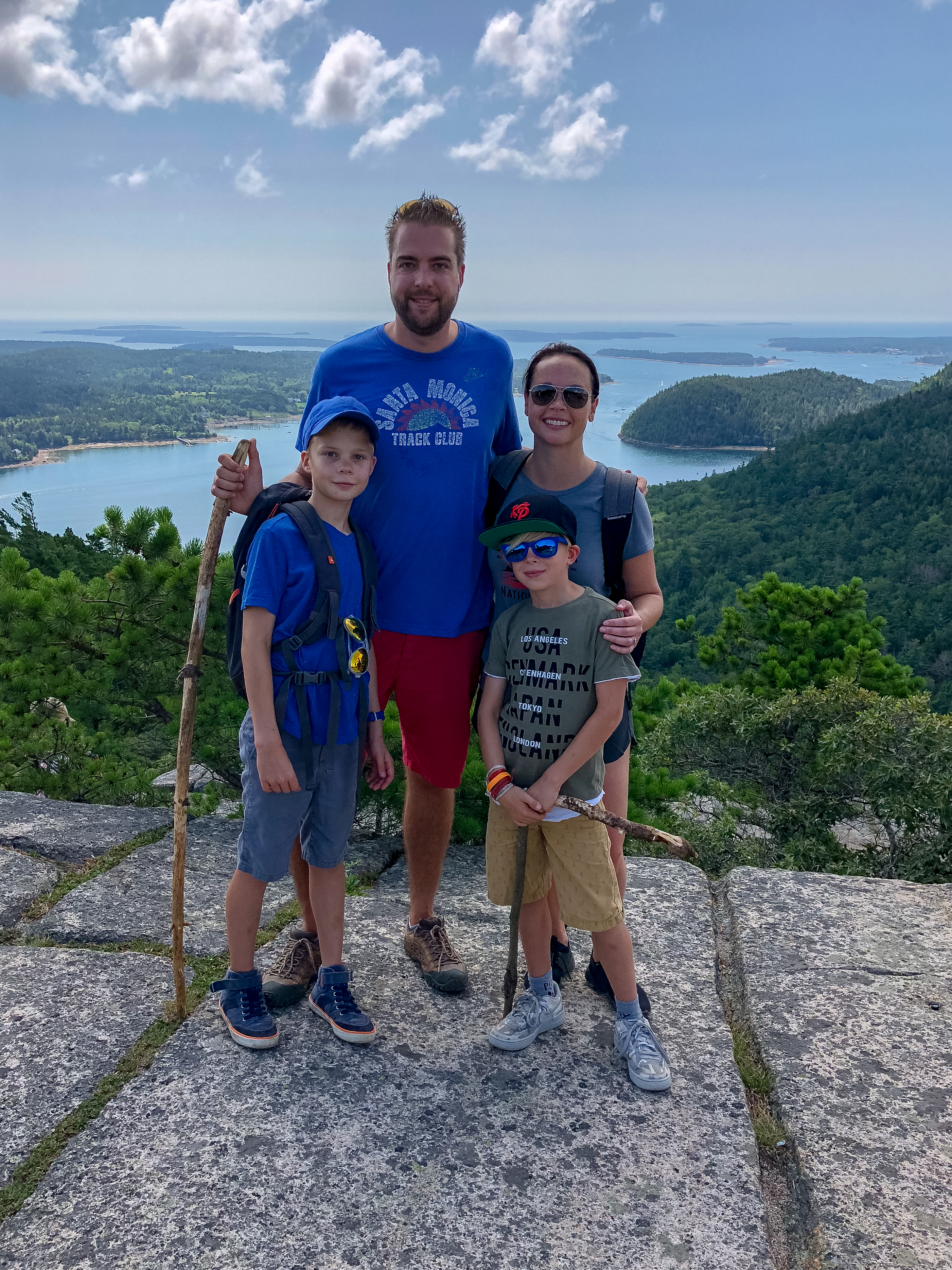 A family of four stands on a rocky ledge, smiling after a hike, holding walking sticks.