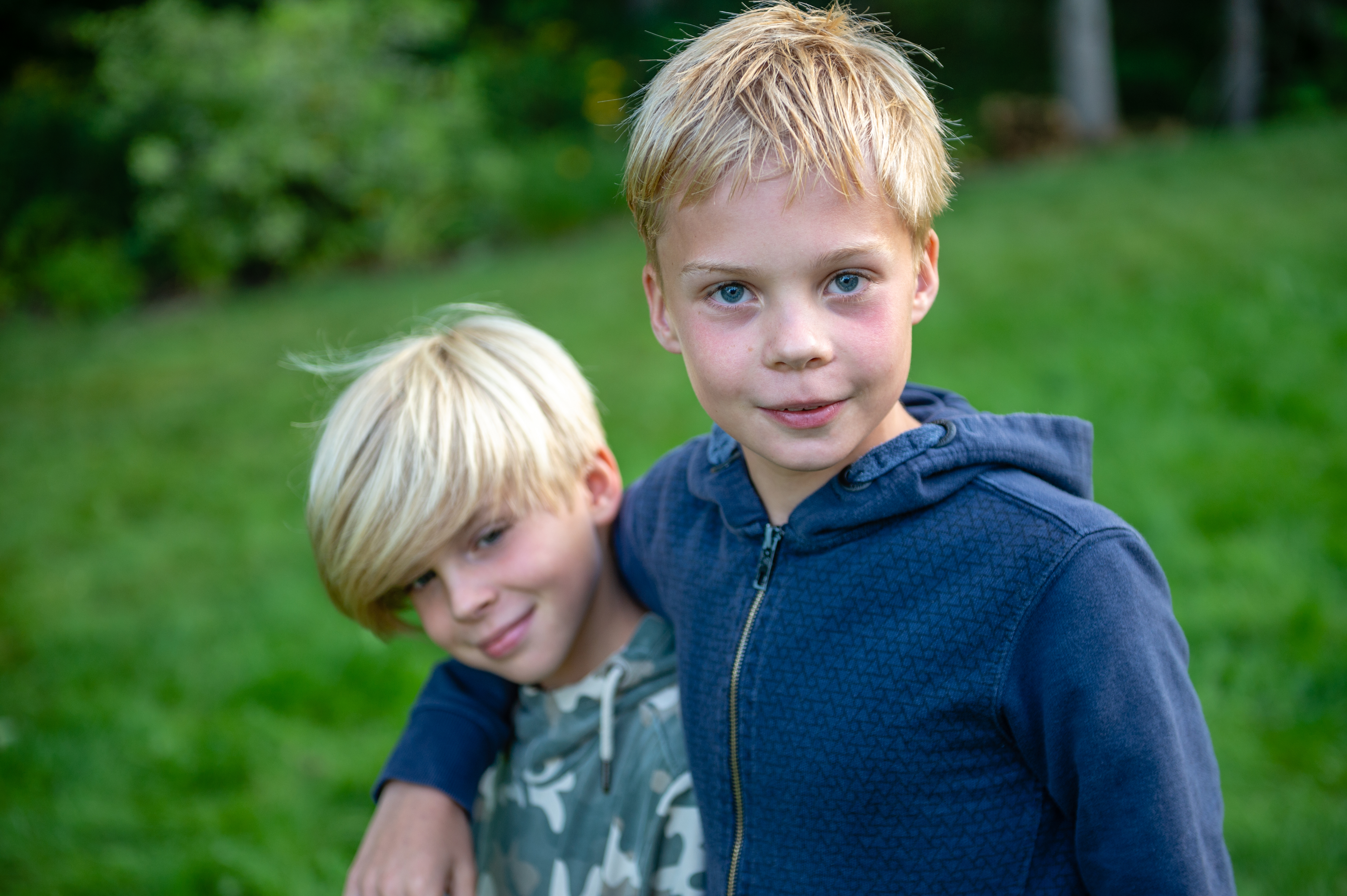 Two young boys stand close together outdoors, one with his arm around the other, both smiling at the camera.