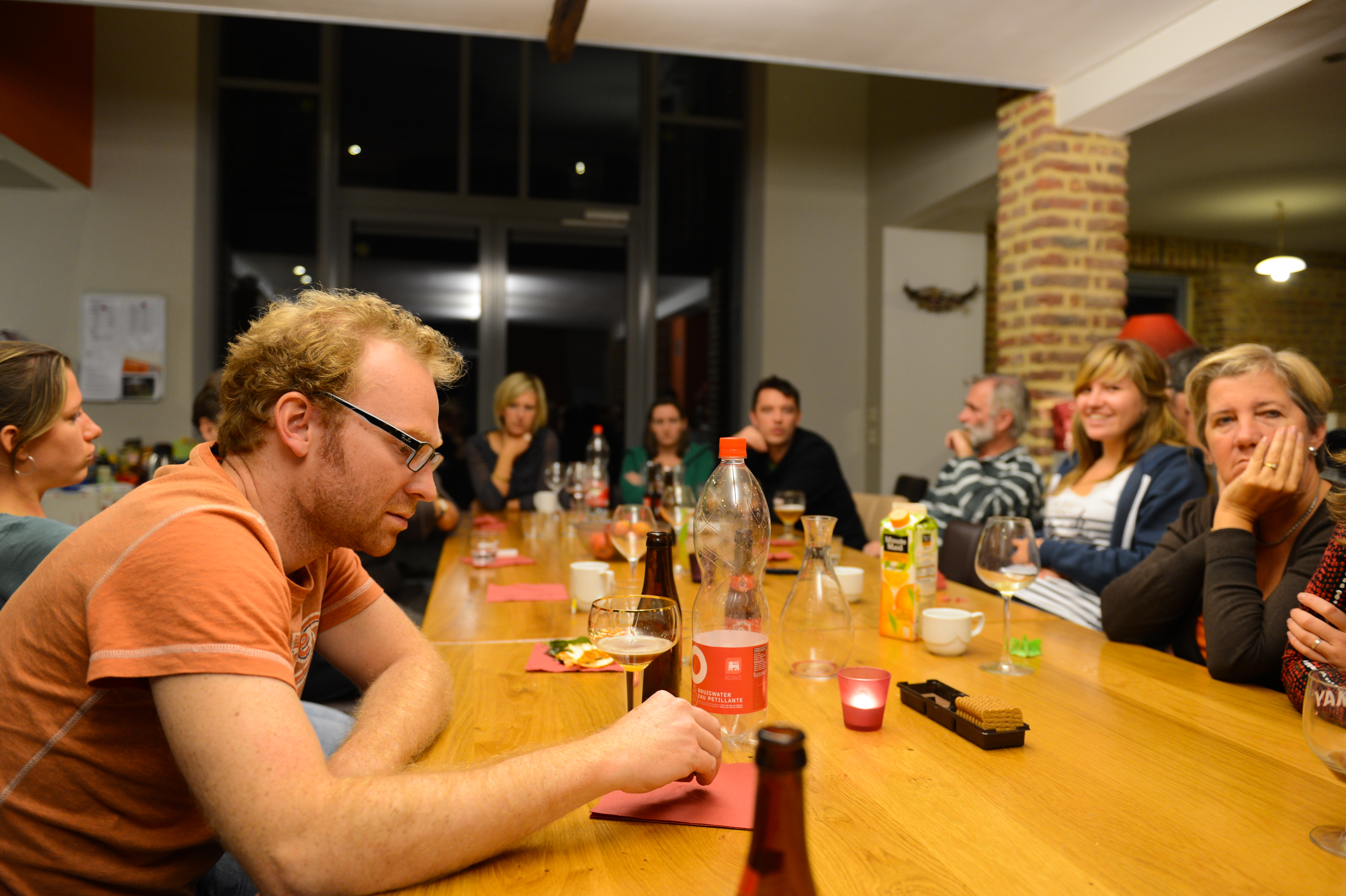 A man in glasses sits at a table with drinks and food, surrounded by a group of people socializing.