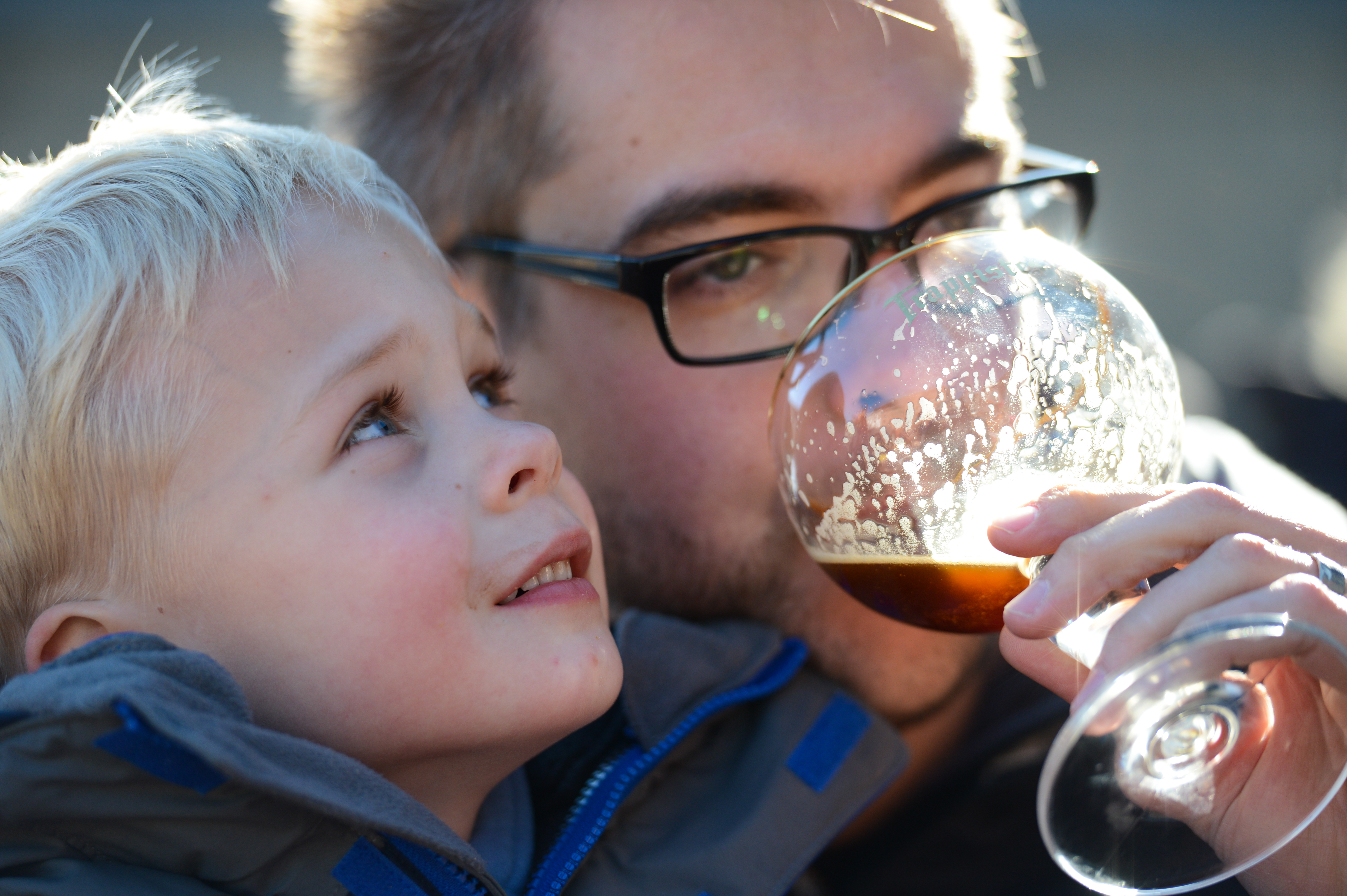 A man wearing glasses drinks from a glass of dark beer while a young child looks up at him.