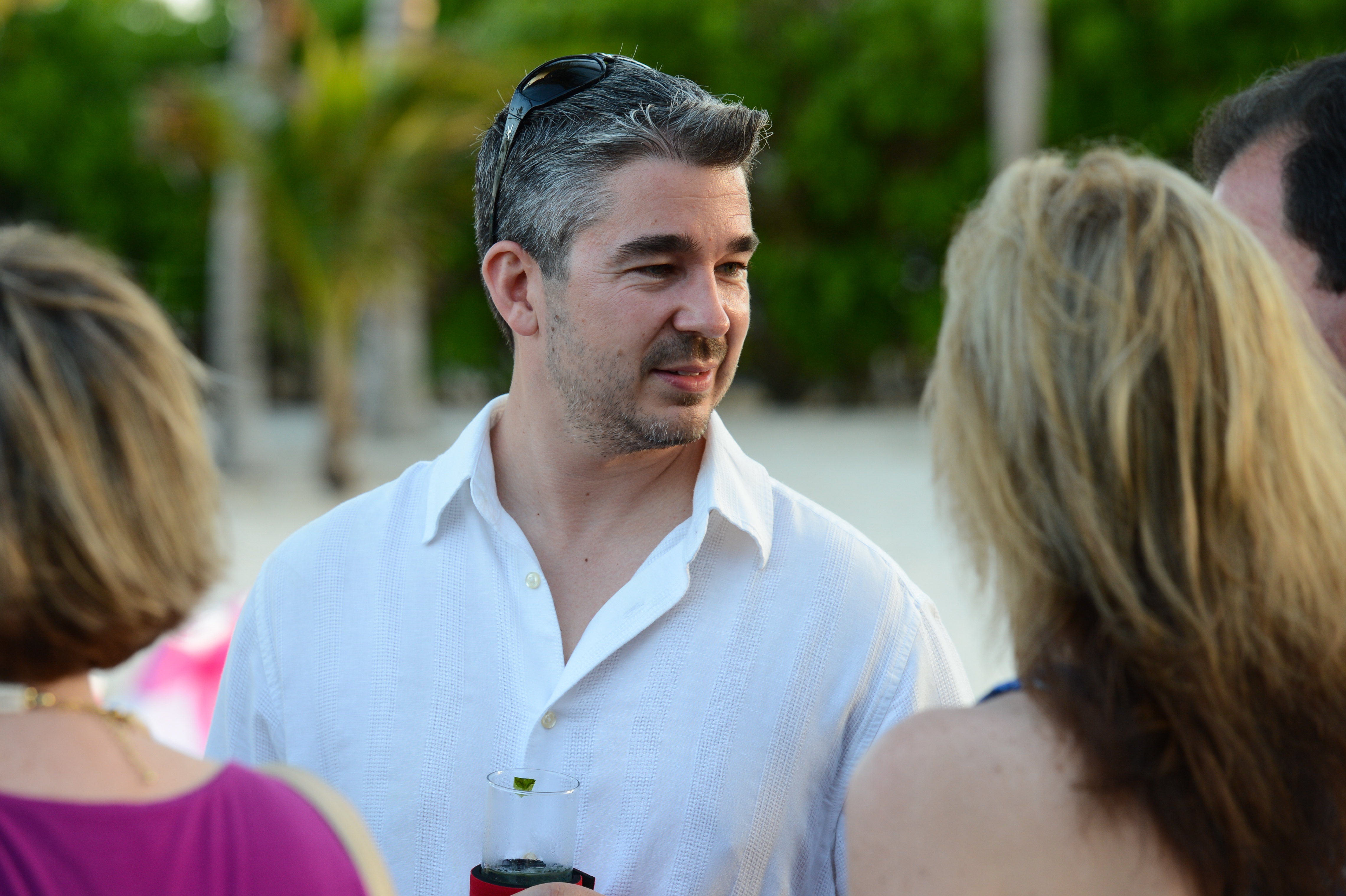 A man in a white shirt holds a drink and talks with three people at an outdoor gathering.