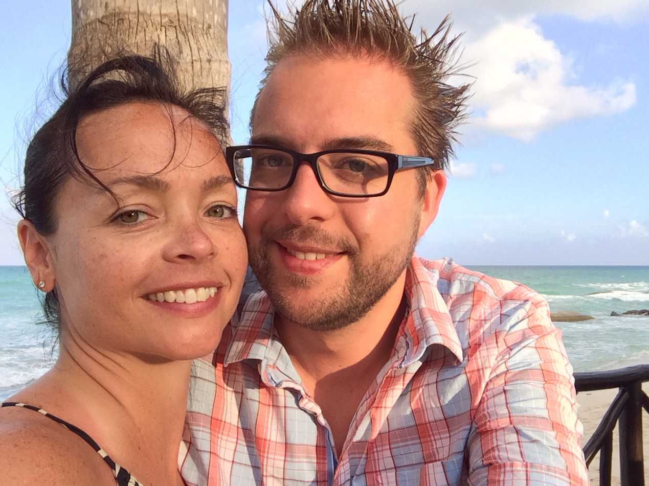 A smiling couple takes a close-up selfie on a beach in Cancun, with the ocean in the background.