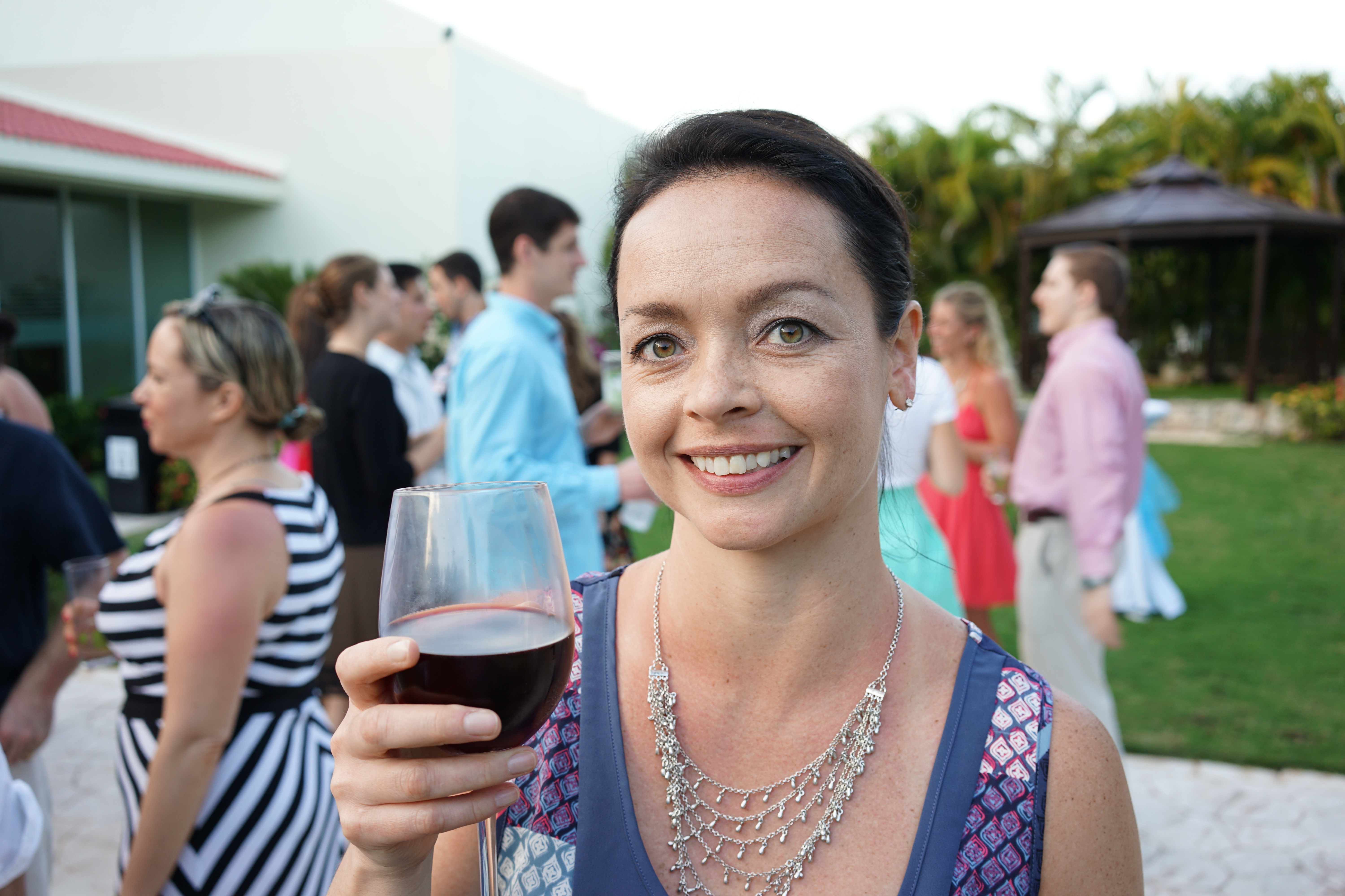 A smiling woman holds a glass of red wine at an outdoor award party with other guests in the background.
