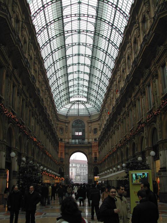 A crowded shopping gallery in Milan with a glass ceiling, decorated with lights and people walking through.
