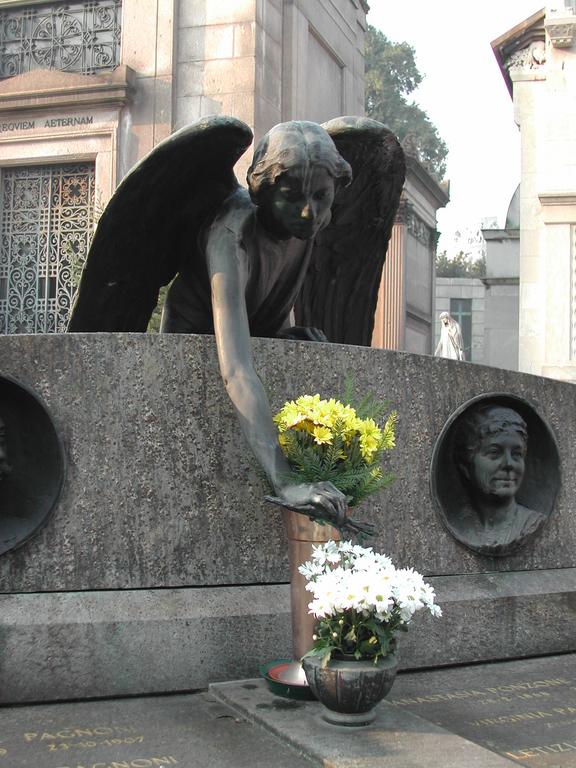 A bronze angel statue leans forward, reaching toward a vase of yellow flowers on a stone grave monument.