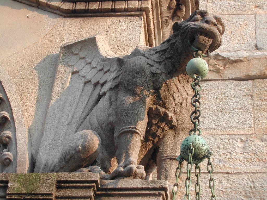 Stone gargoyle with wings holding a chain with metal spheres in its mouth, mounted on a historic building.