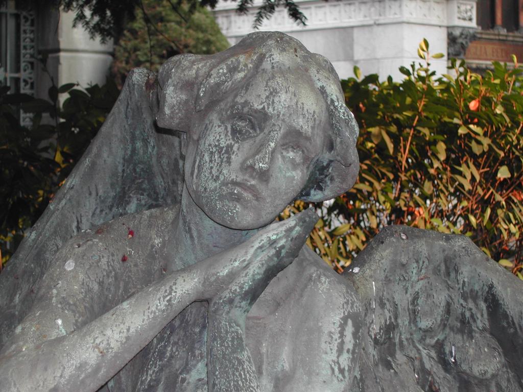 A weathered bronze statue of an angel with a sorrowful expression, resting its head on crossed hands.