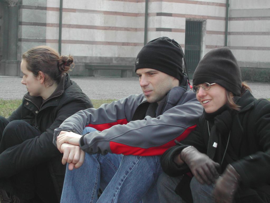 Three people in warm clothing sit on the ground, two smiling while one looks away.