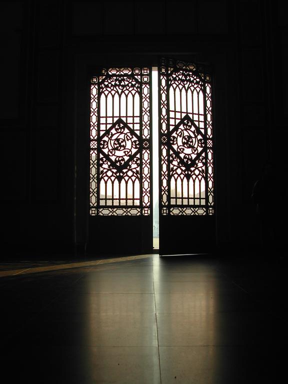 Sunlight shines through ornate metal doors, casting shadows on the floor in a dimly lit interior.