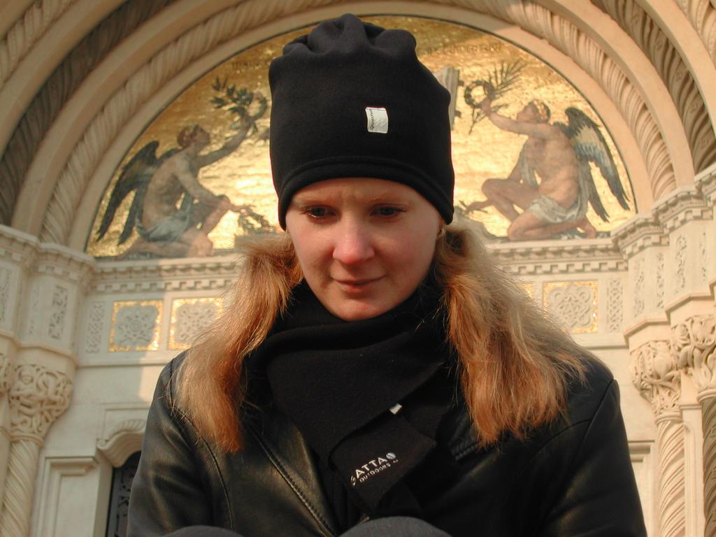 A woman in a black hat and jacket looks down, sitting in front of an ornate building with golden artwork.