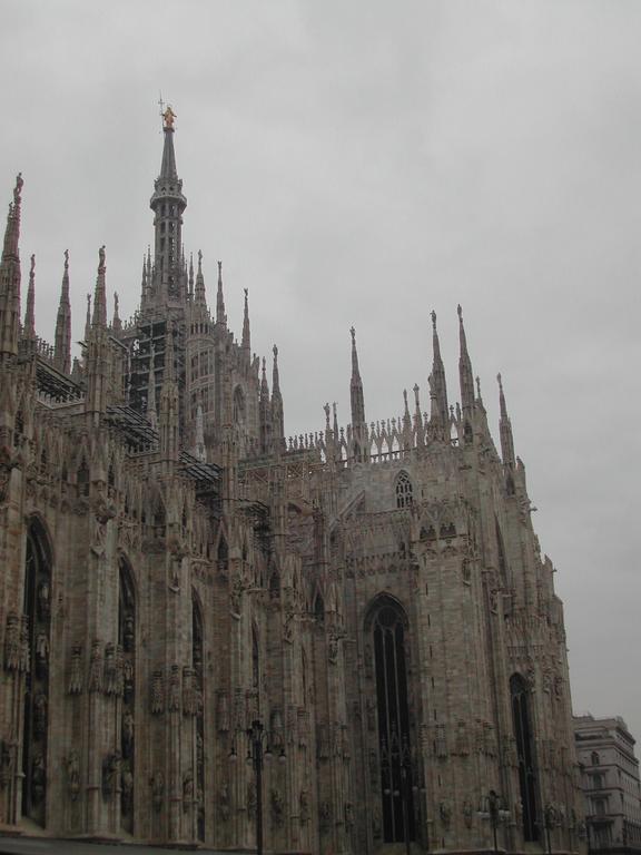 The Milan Cathedral's detailed Gothic facade with tall spires and statues, set against a cloudy sky.