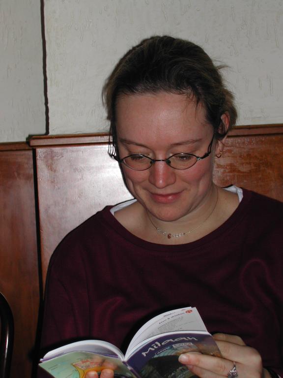 A woman wearing glasses and a burgundy sweater sits indoors, reading a travel guidebook about Milan.