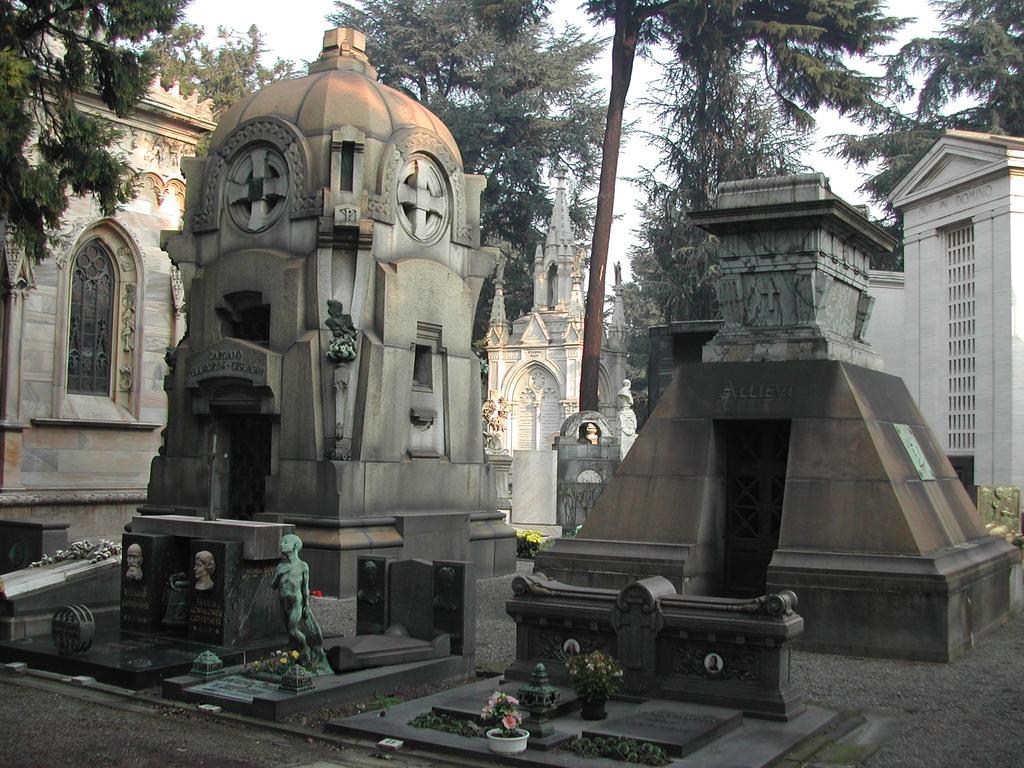 A cemetery in Milan with elaborate stone mausoleums, statues, and tombs surrounded by trees.