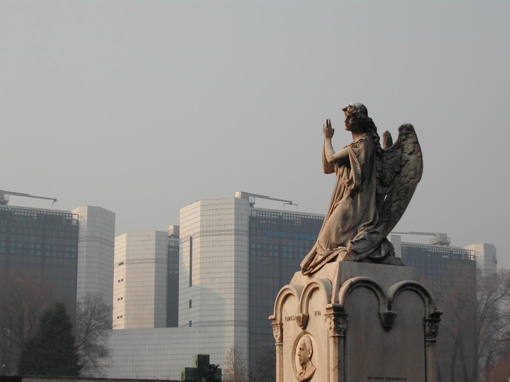 A stone angel statue on a monument with modern glass buildings in the background.