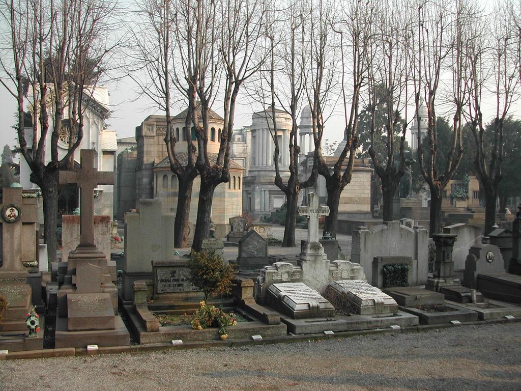 A cemetery with tombstones, crosses, and leafless trees, with large mausoleums in the background.