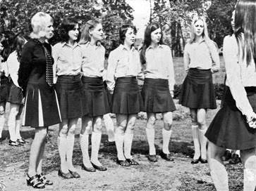 A group of young women in uniforms stands in a line outdoors, listening to another woman addressing them.