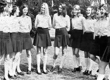 A group of young women in matching skirts and blouses stand outside, smiling and talking together.