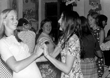 Two young women smile and hold hands while dancing at a lively indoor gathering with others in the background.