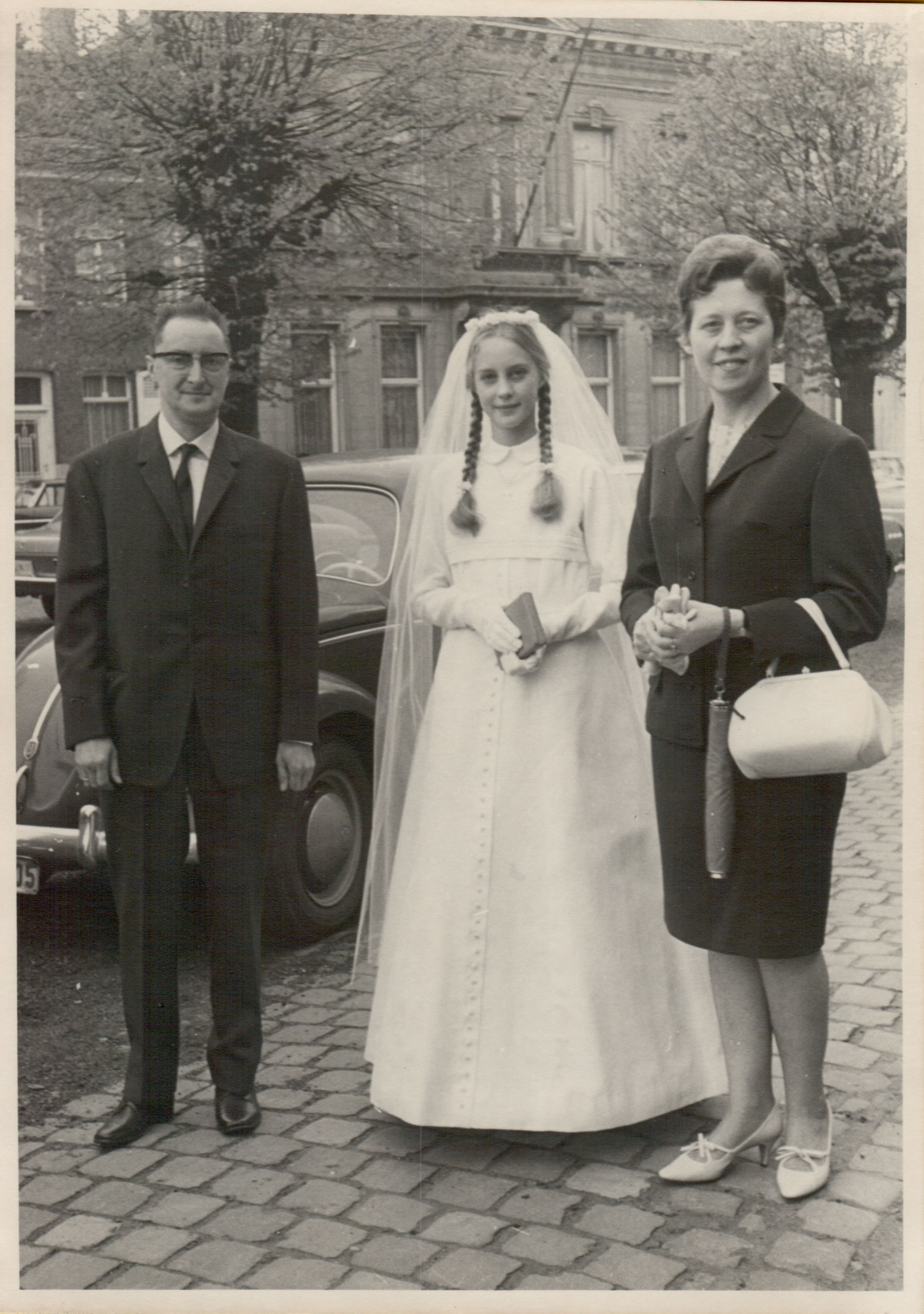A young girl in a white dress and veil stands between two adults, holding a small book and smiling.
