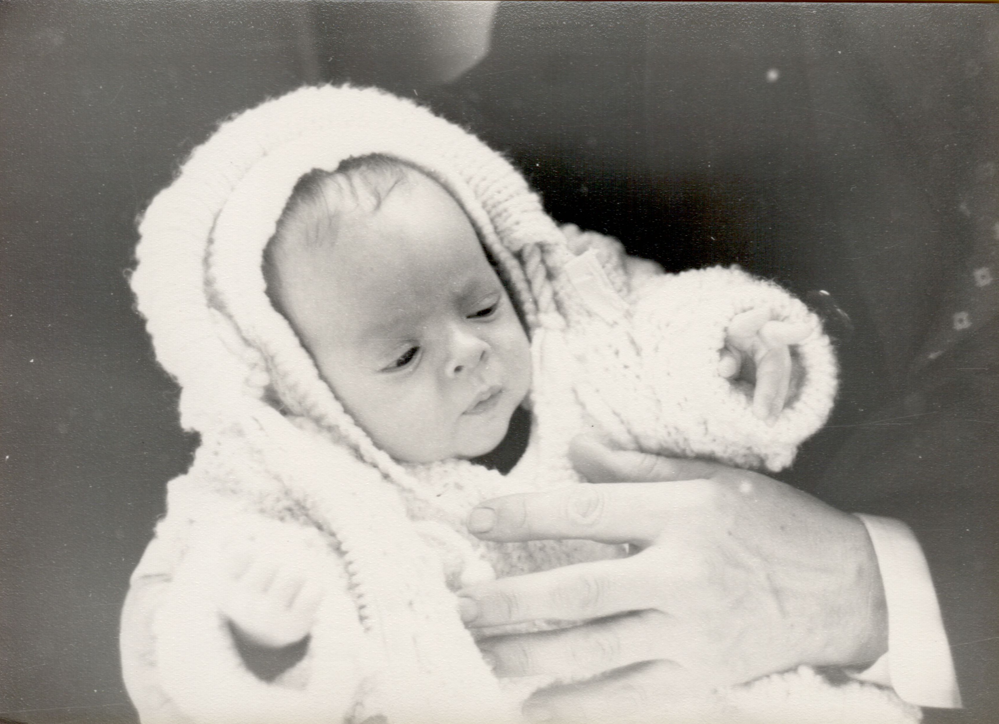 A baby dressed in a knitted outfit is held by an adult during a baptism ceremony.