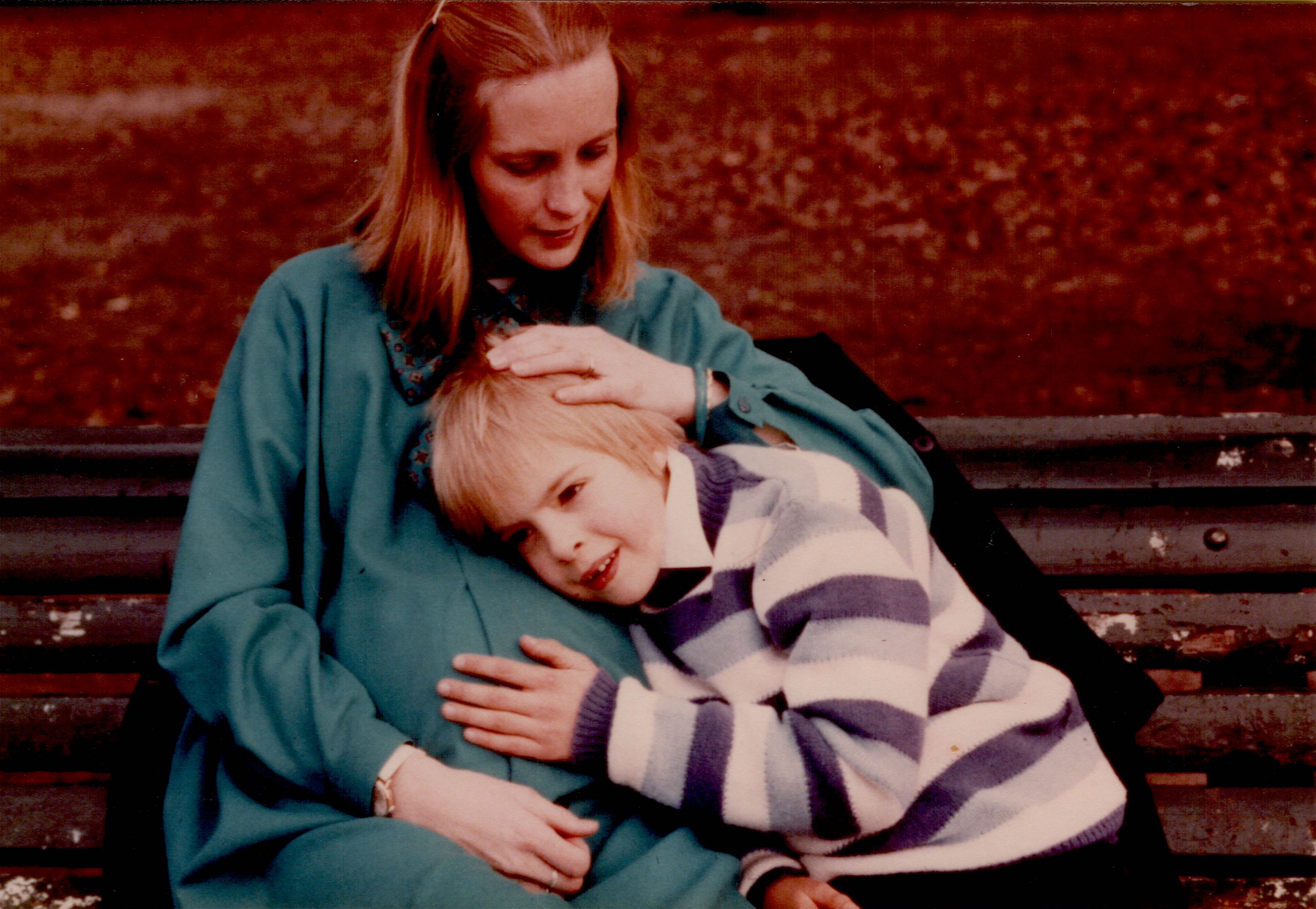 A pregnant mother sits on a bench while her child rests their head on her belly, smiling.