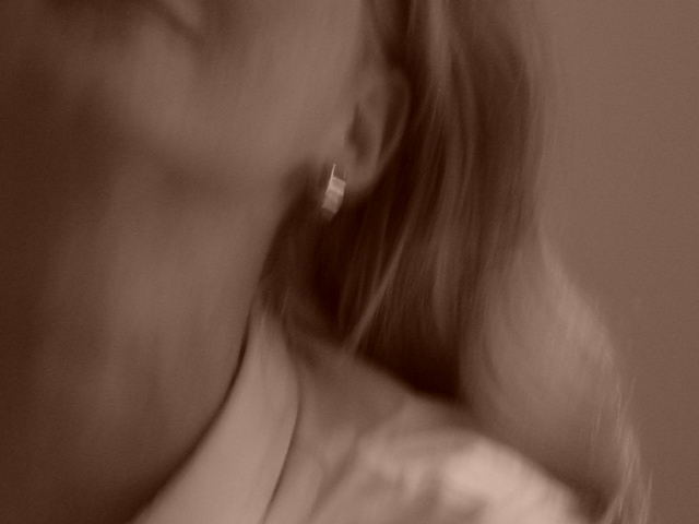 Blurry close-up of a person with light hair, wearing a white shirt and an earring, tilting their head.