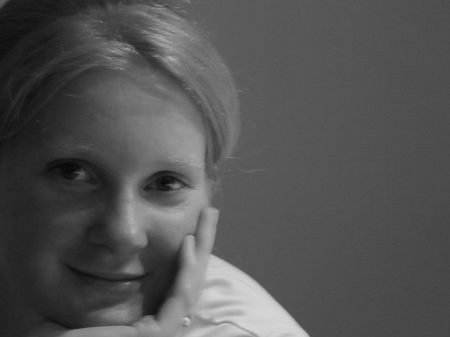 A person with light hair smiles gently, resting their face on their hand in a black-and-white portrait.