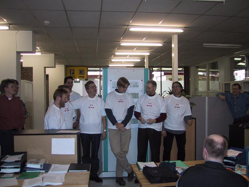 A group of people wearing matching white t-shirts stand in an office, engaged in conversation and presentation.