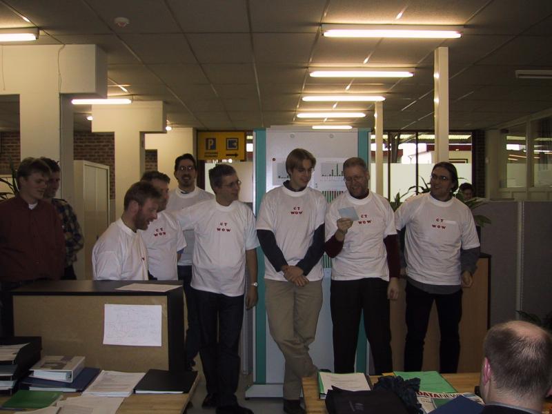 A group of people wearing matching white t-shirts stand in an office, with one person reading from a paper.